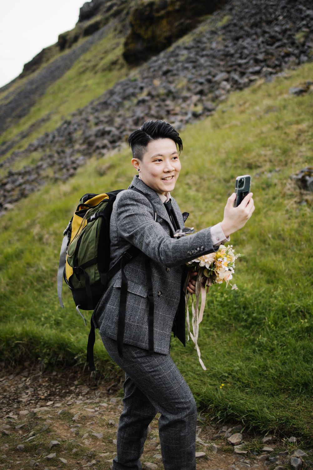 A person in a gray suit carries a backpack while taking photos on a scenic hiking trail.
