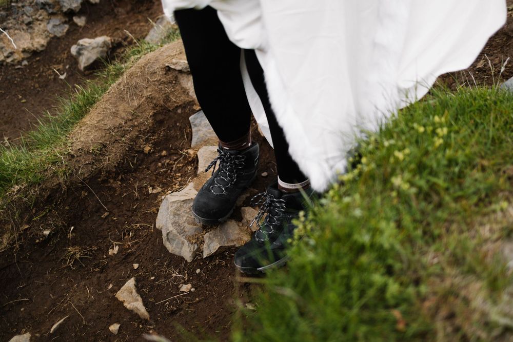 Close-up of hiking boots and a white wedding dress hem on rocky terrain.