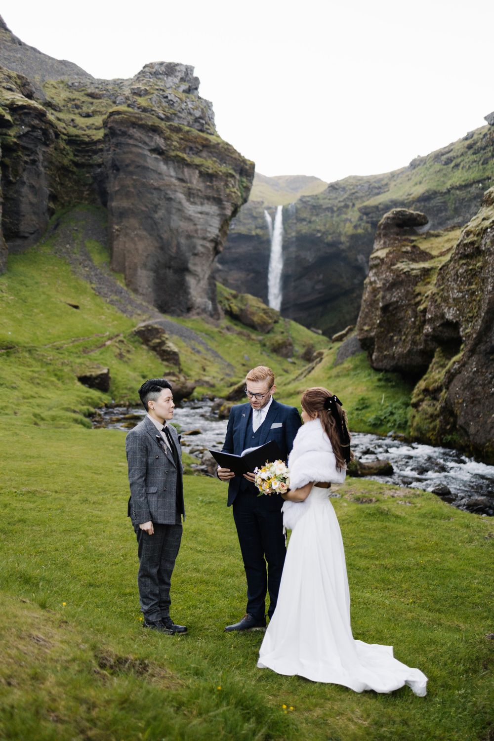 An intimate wedding ceremony takes place in front of a majestic waterfall in Iceland's green landscape.