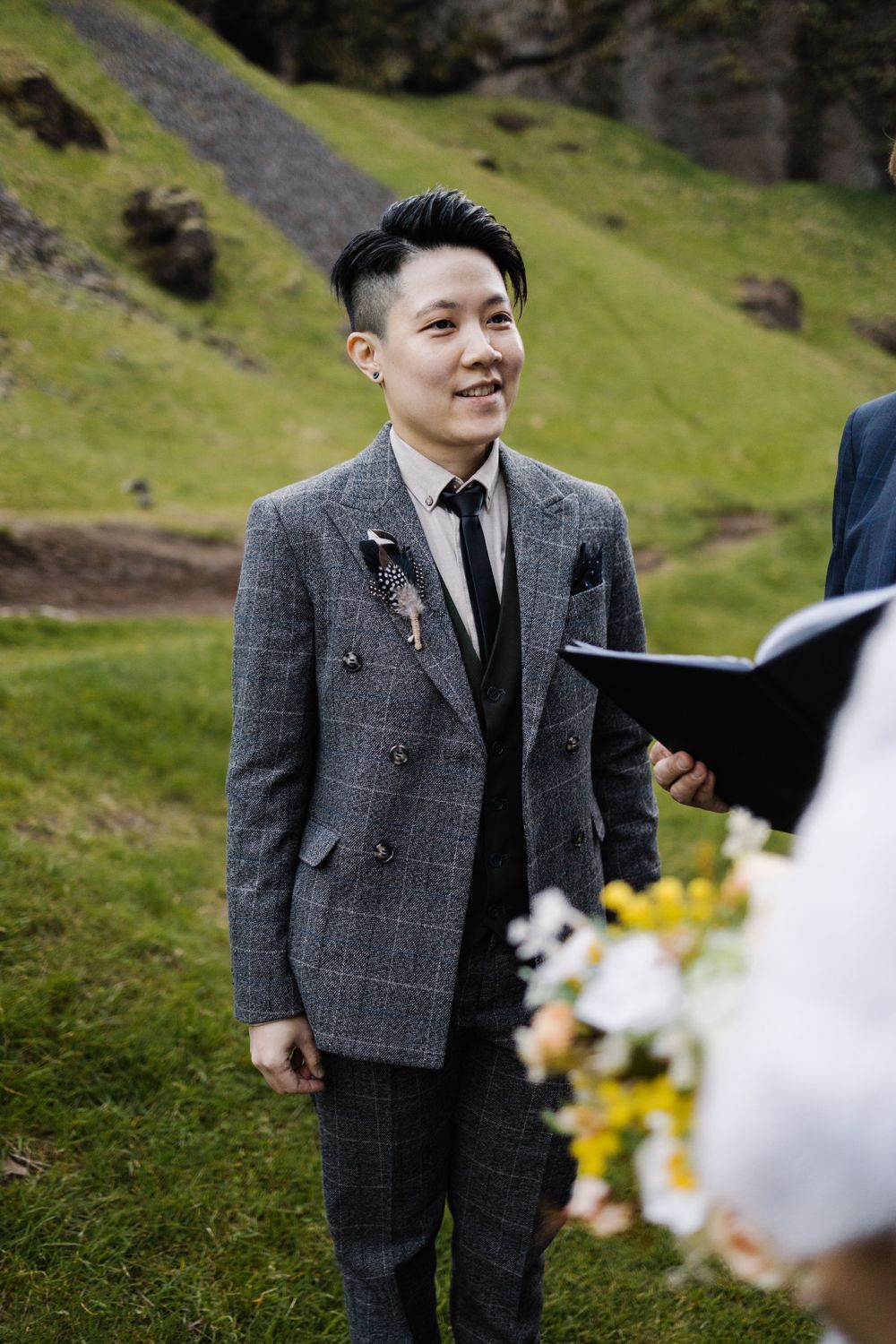 A person in a gray plaid suit and black tie stands on a grassy hillside during an outdoor wedding ceremony.