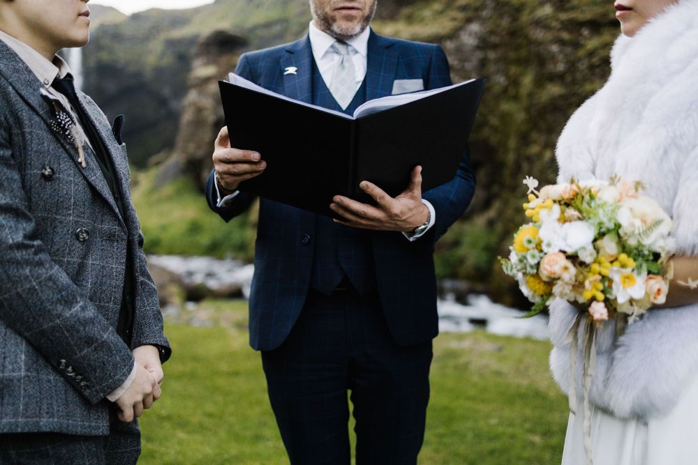 Intimate outdoor wedding ceremony with officiant reading from black book as couple exchanges vows.