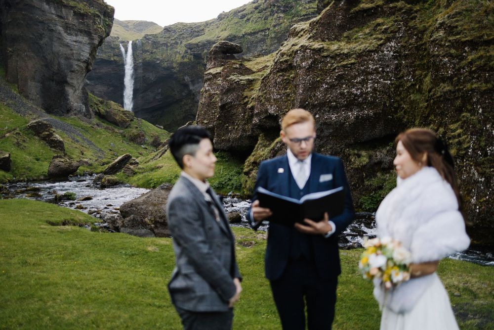 Wedding ceremony taking place near dramatic waterfall with officiant and couple in natural setting.