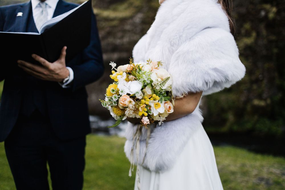 Detail shot of wedding ceremony with white fur wrap and yellow bouquet against natural backdrop.