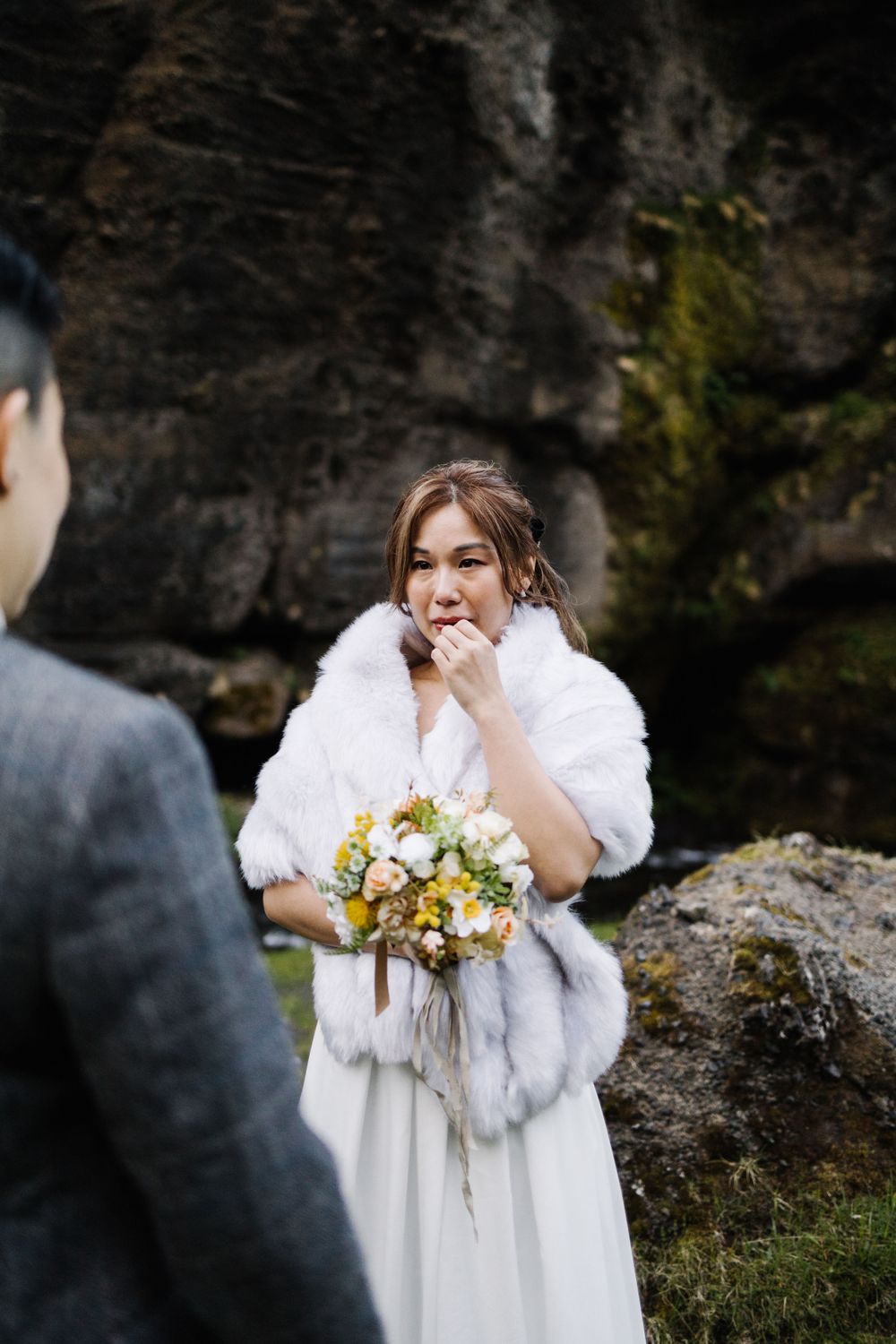Intimate outdoor wedding ceremony in front of dramatic rocky cliffs with white fur wrap and bouquet.