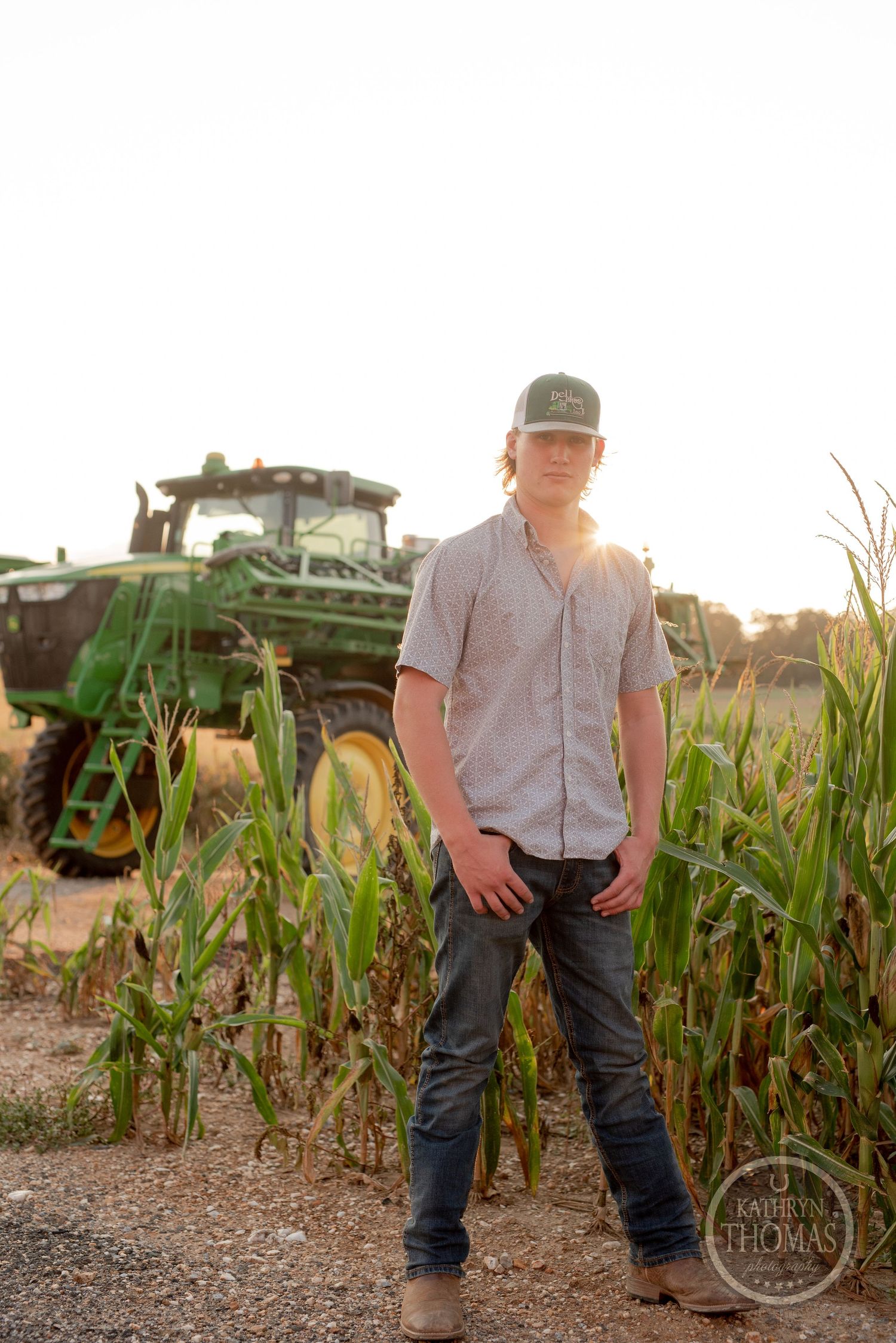 Farmer poses beside cornfield with John Deere tractor in background during sunset.