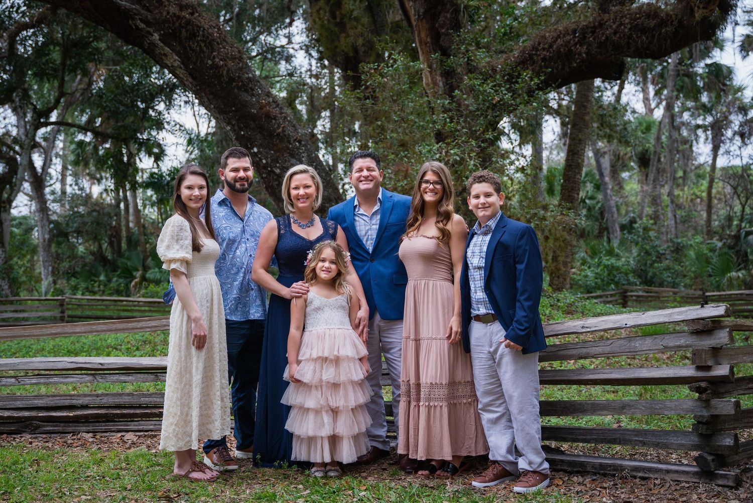 Formal family group photo in front of rustic wooden fence and lush trees.