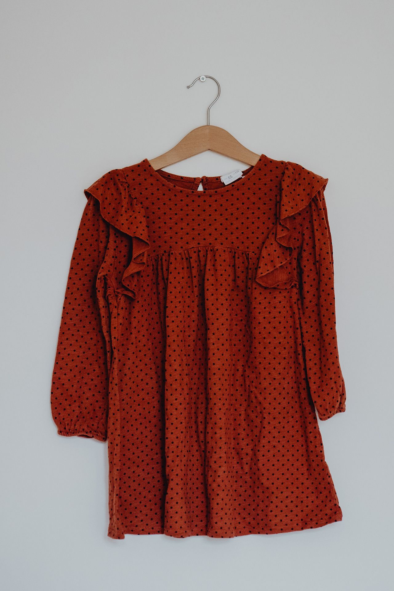 A red polka dot dress with ruffled shoulders hangs on a wooden hanger against a white wall.
