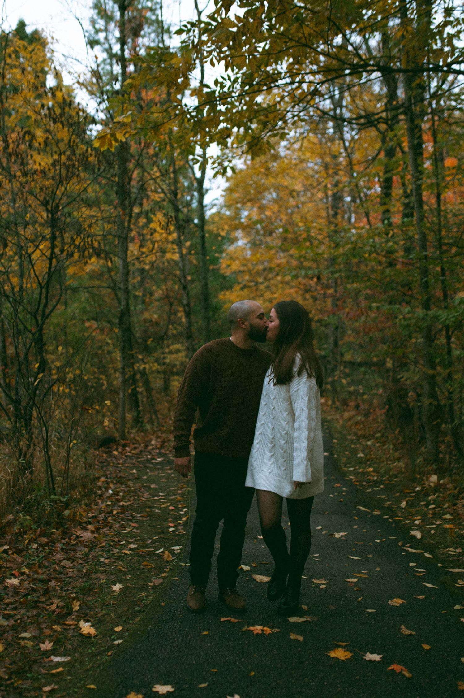 A couple sharing a kiss on a leaf-covered forest path in autumn.