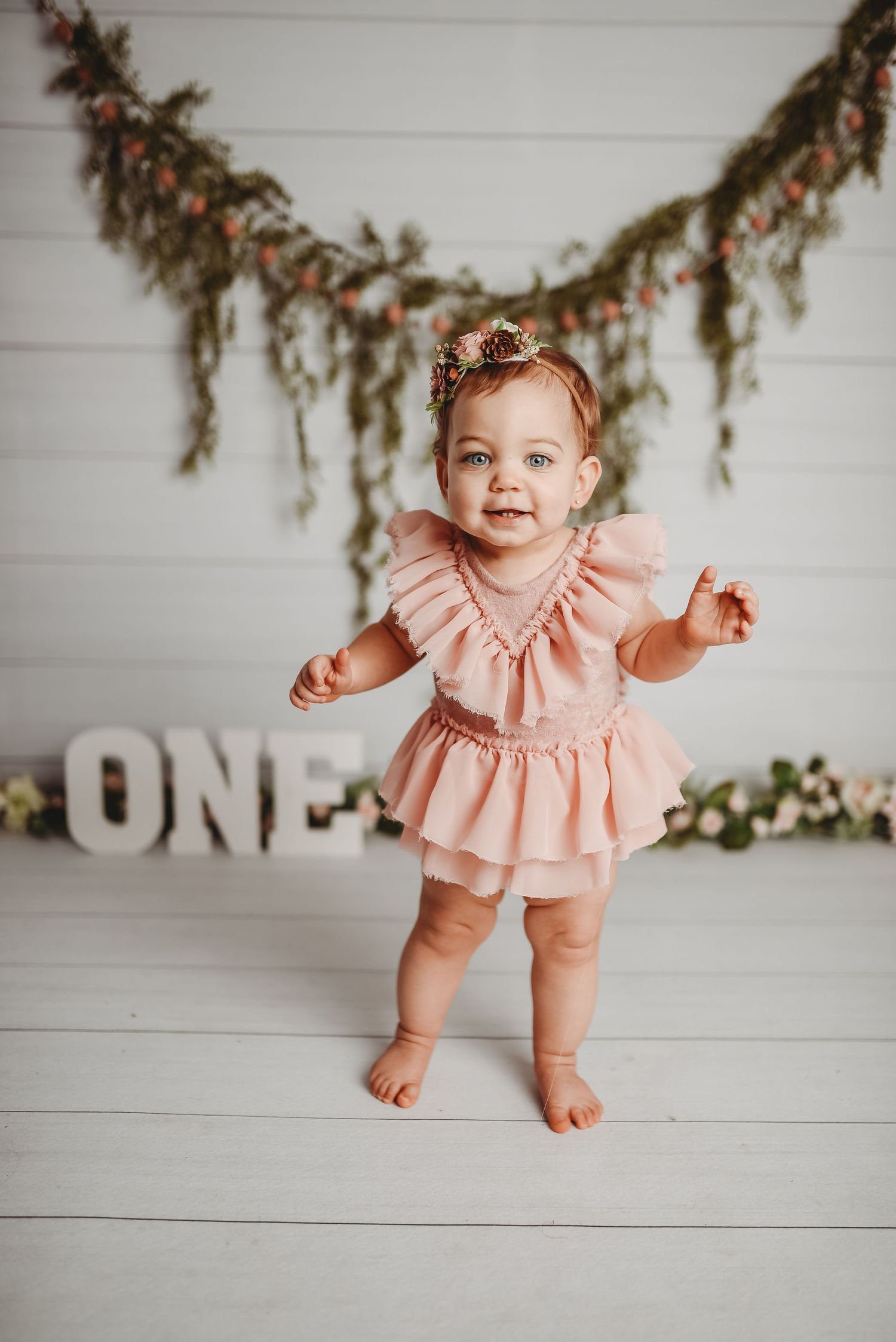 Baby wearing a peach-colored ruffled outfit sits on white wooden floor with decorative greenery.