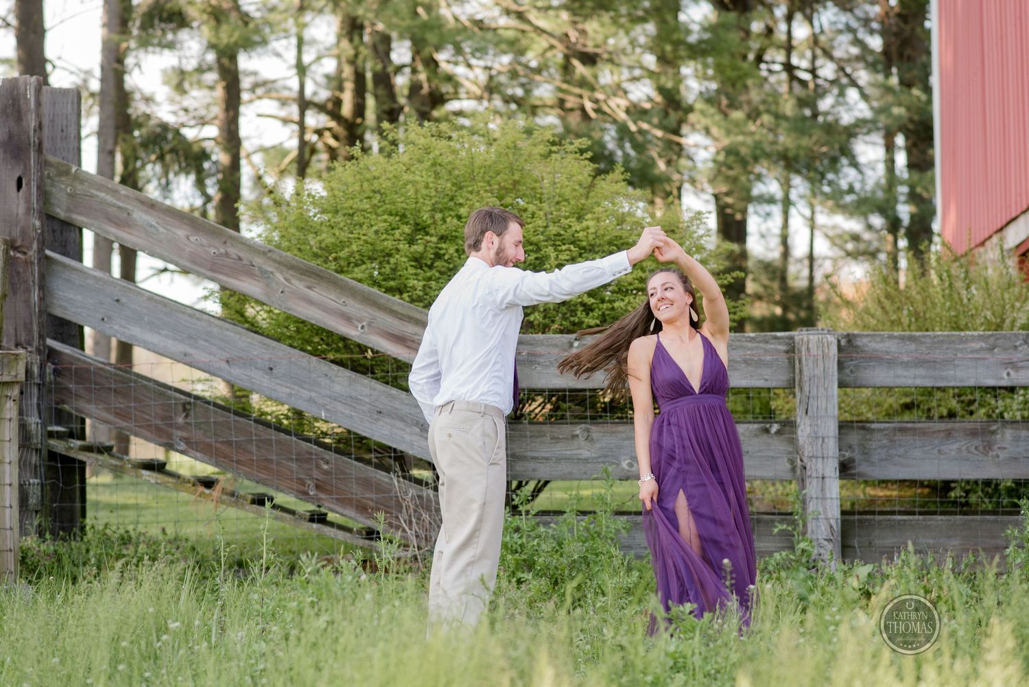 Couple dancing outdoors by wooden fence, woman in flowing purple dress and man in white shirt.