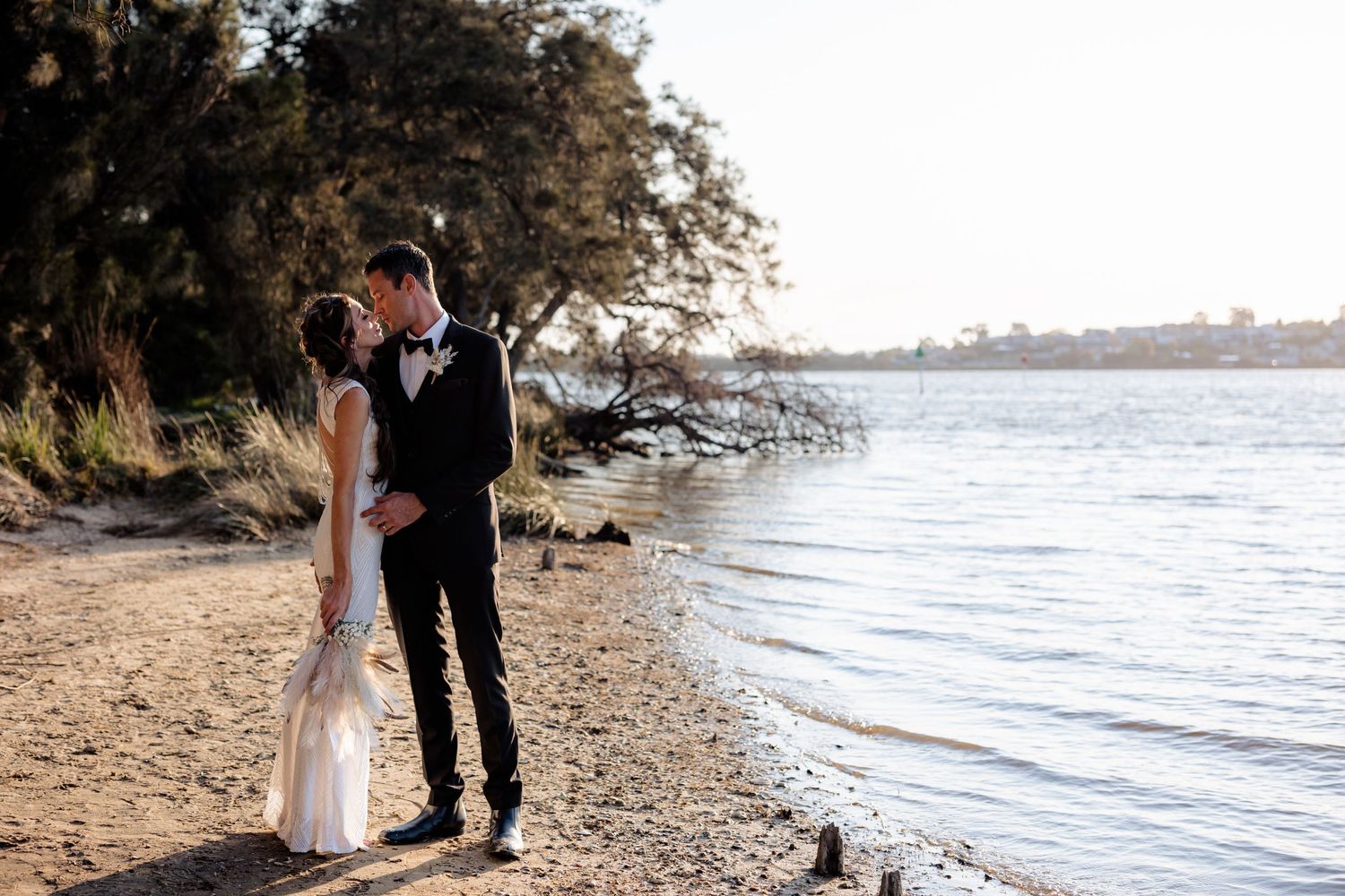 Couple embracing on a scenic beach shoreline with water and trees in the background.