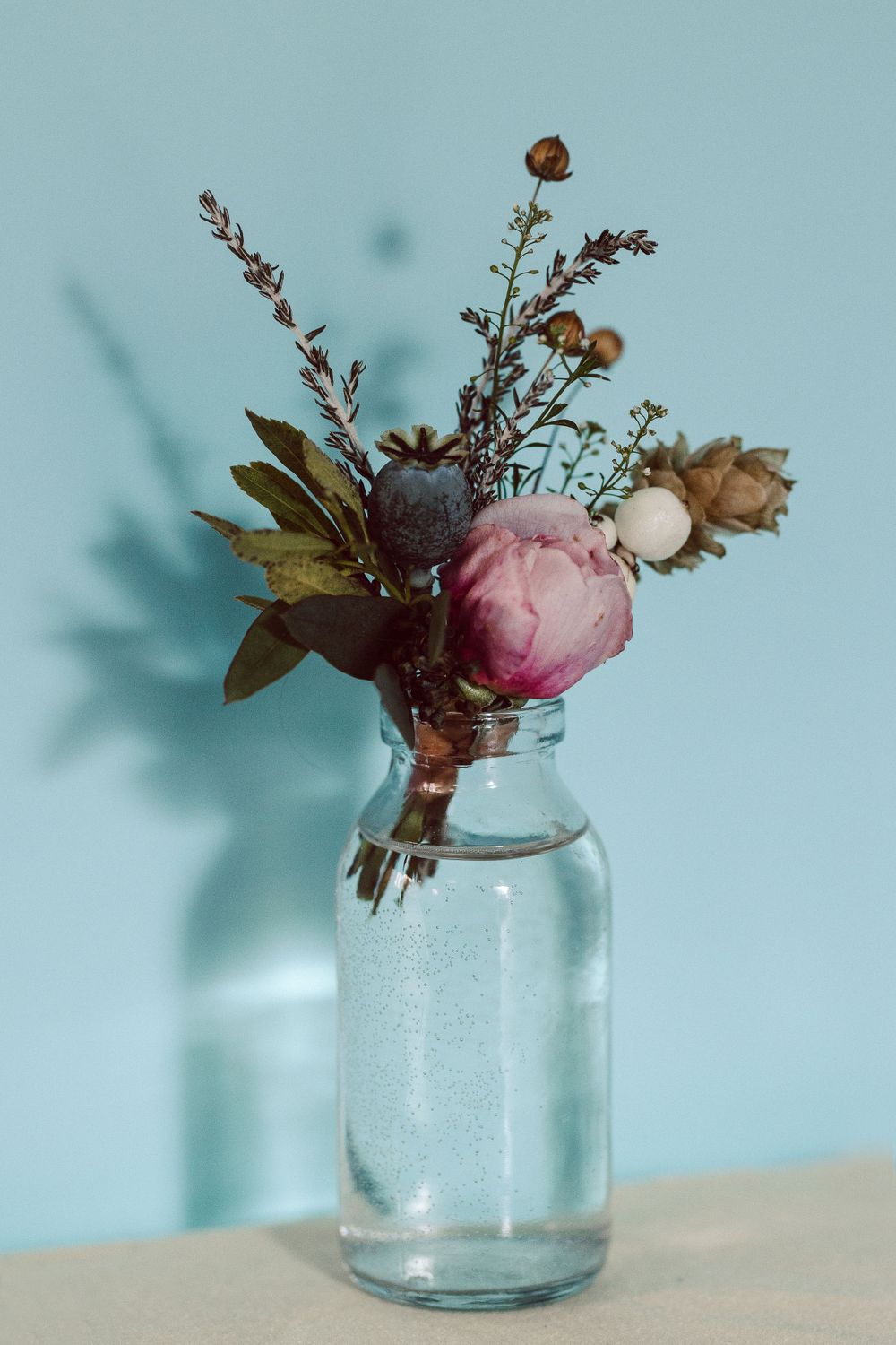 Small glass jar with delicate pink flowers and greenery against light blue background.