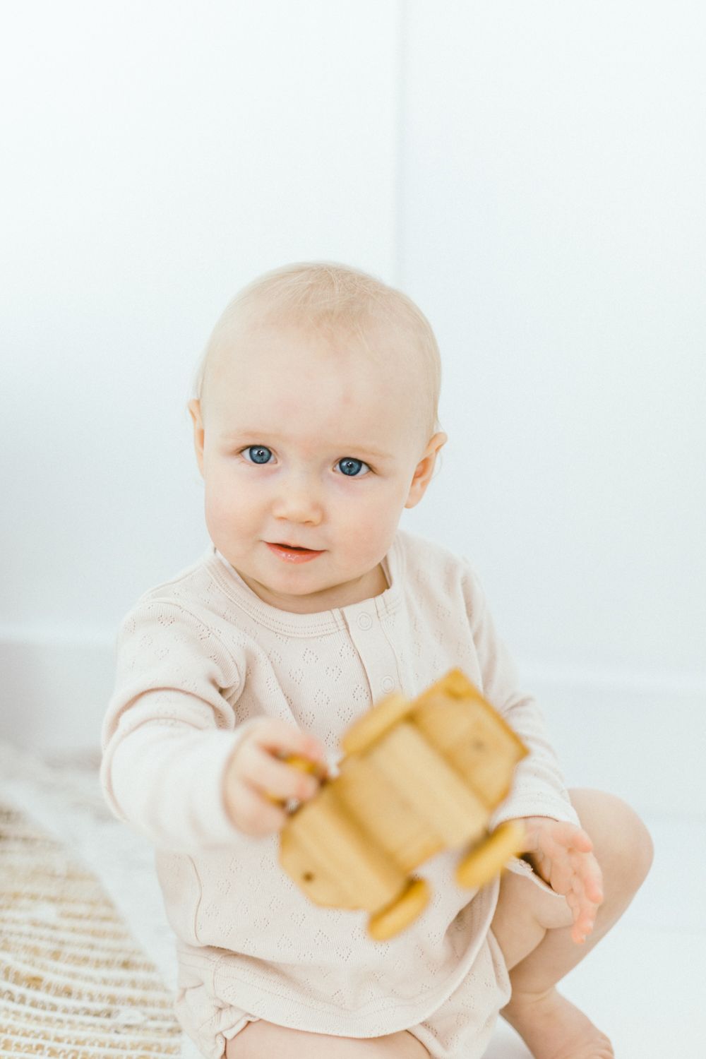 Baby in light colored outfit holding wooden toy against white background.