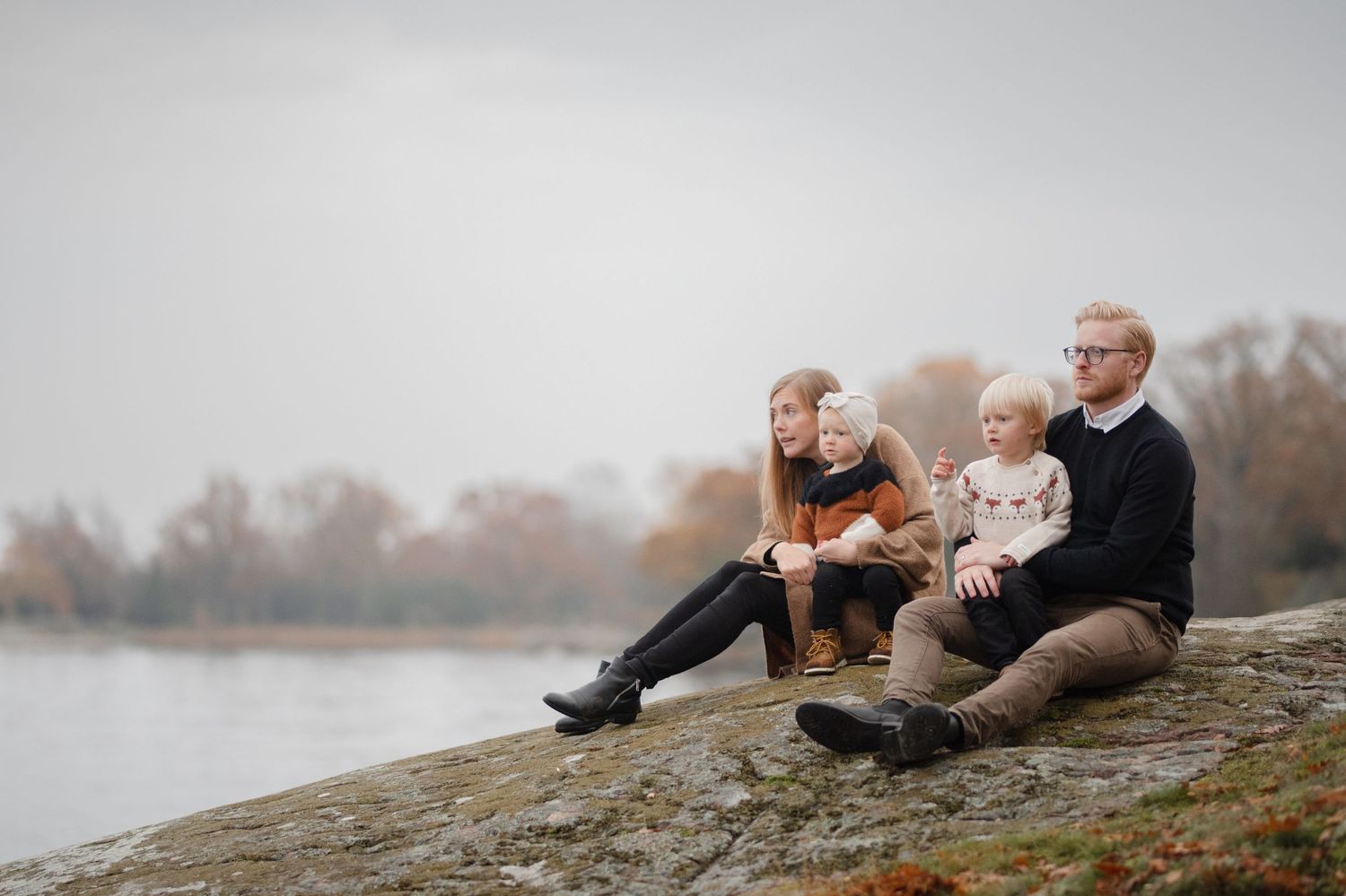 Family sitting together on a rocky outcrop by a peaceful lake on an overcast day.