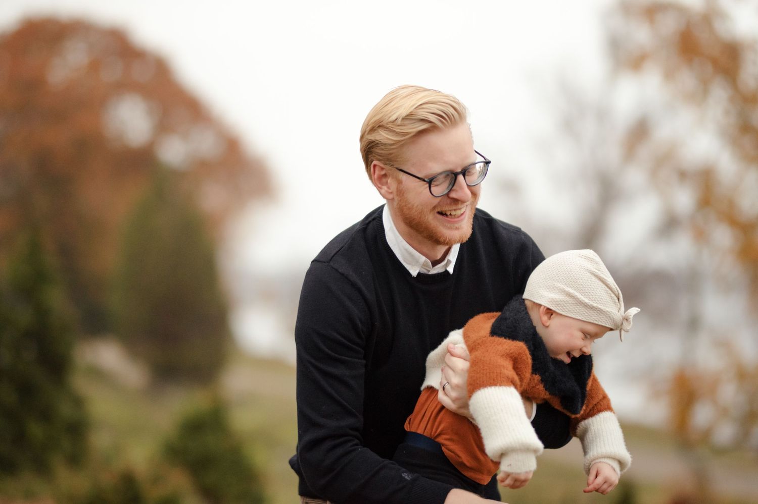 Person in black sweater holding small child wearing winter hat and orange sweater outdoors.