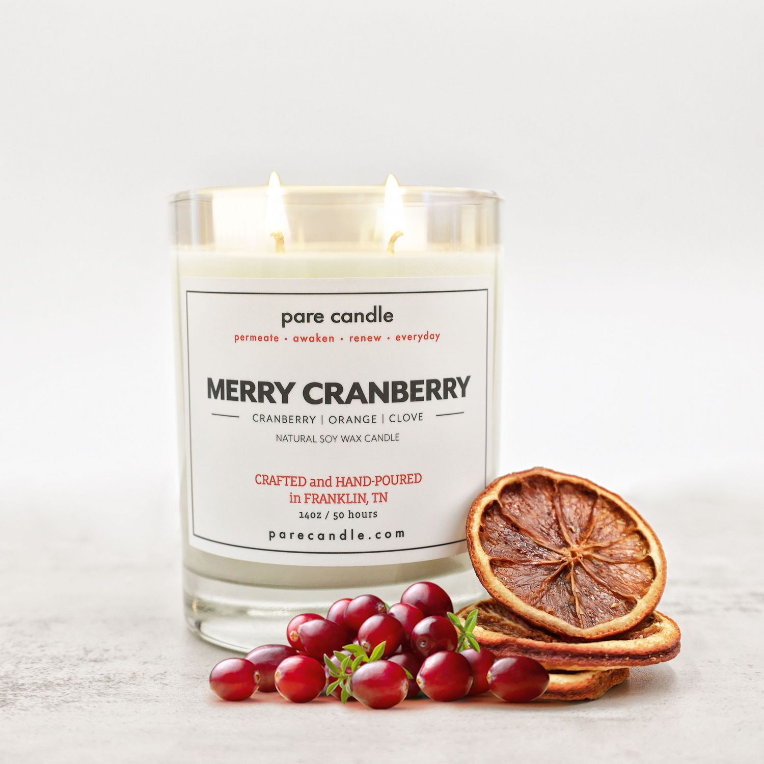 Candle photography. White candle in a glass jar labeled 'Merry Cranberry' with fresh cranberries and dried citrus slices beside it.