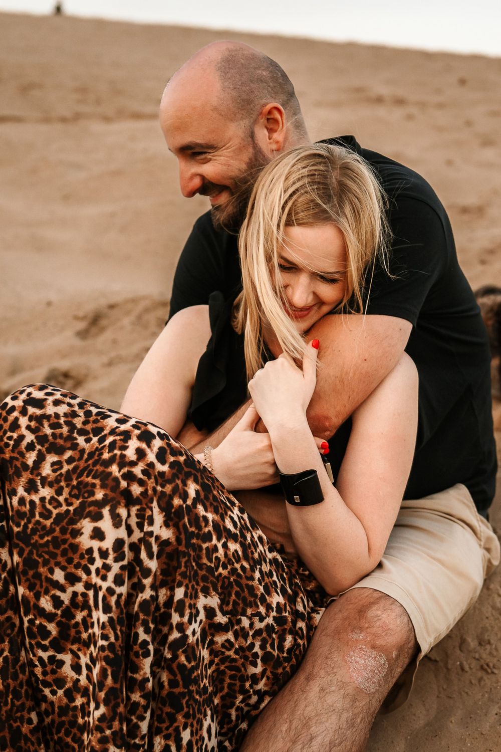Joyful couple shares intimate moment on sandy beach wearing leopard print dress and casual attire.