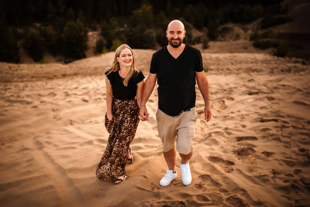 Couple in casual attire smiling while walking on sand dunes during sunset.