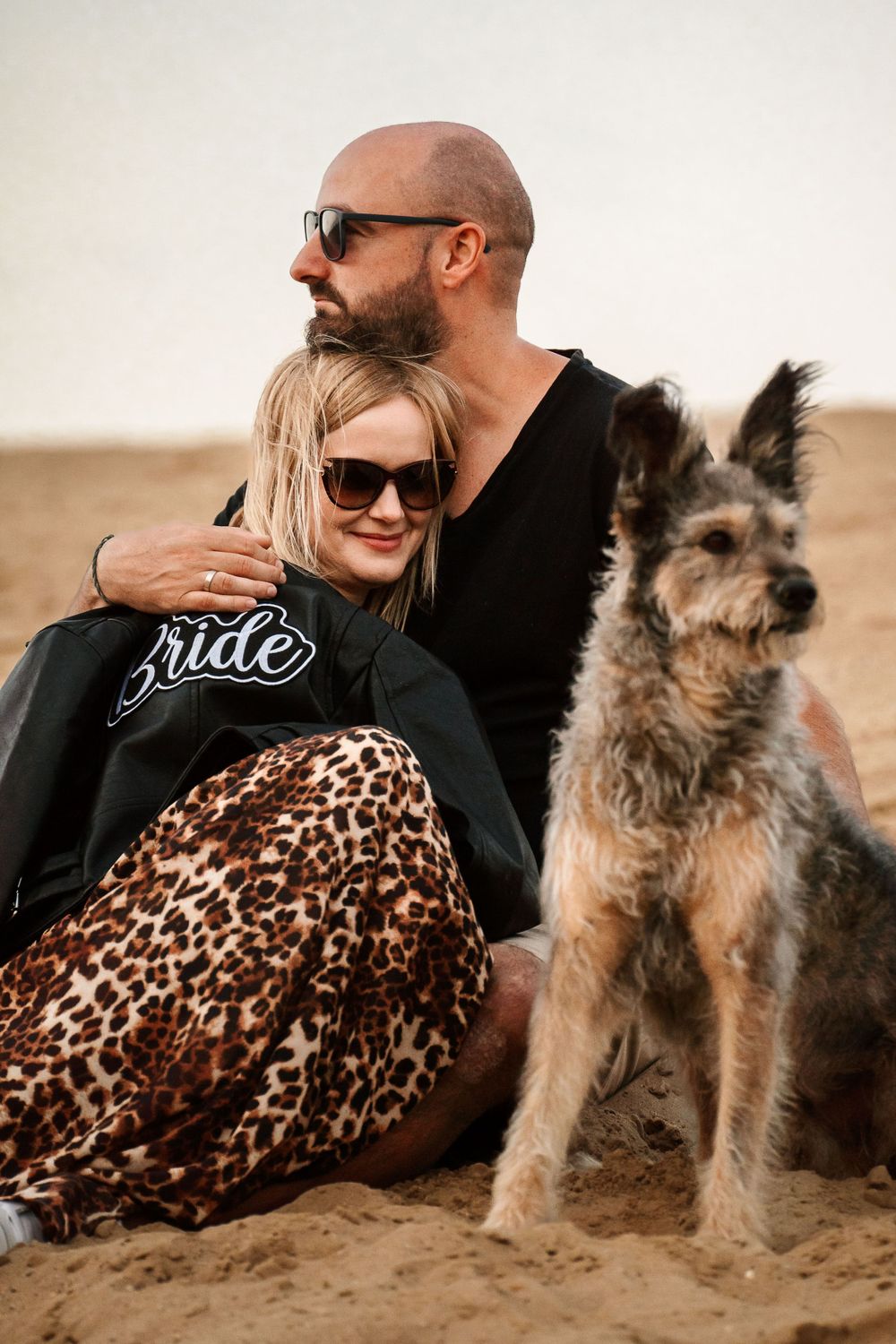 Couple in casual wear sits with their terrier dog on beach wearing sunglasses.