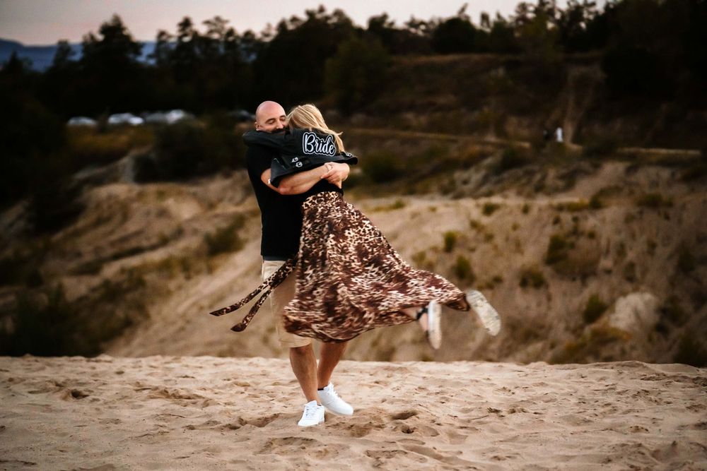 Couple dancing on clifftop during sunset wearing flowing dress and casual attire.