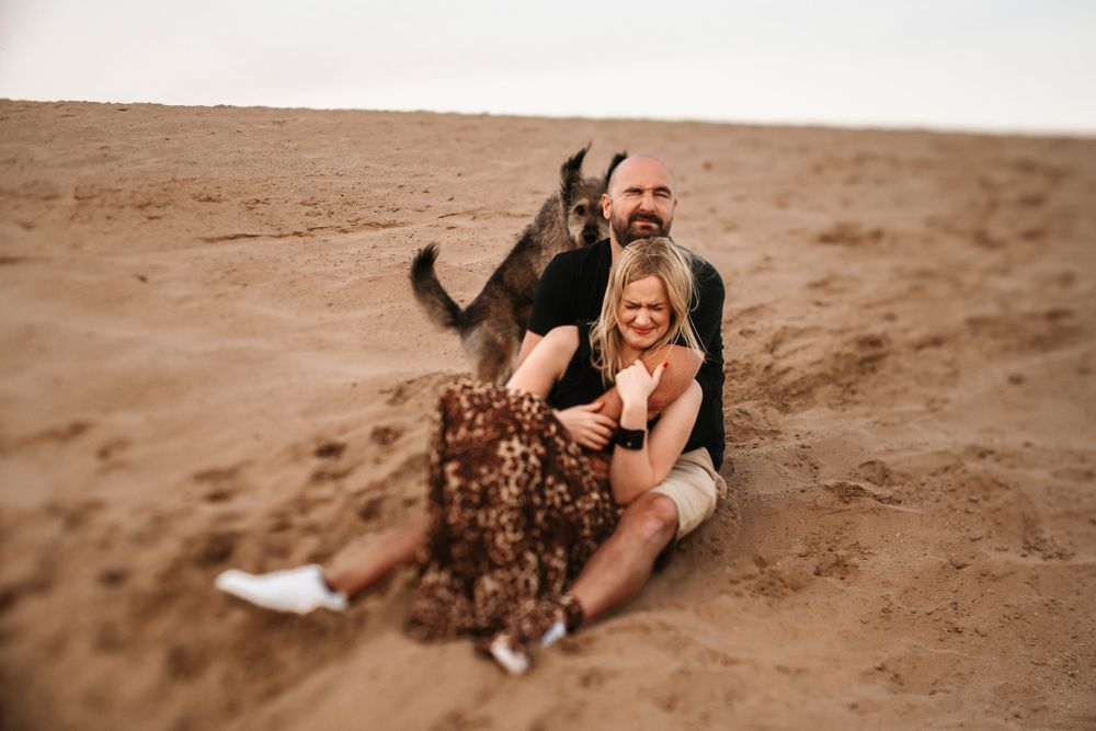 Playful couple lounges on sandy dunes with dog visible in background.