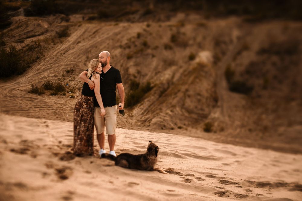 Couple embracing on sandy hillside with dog during sunset.