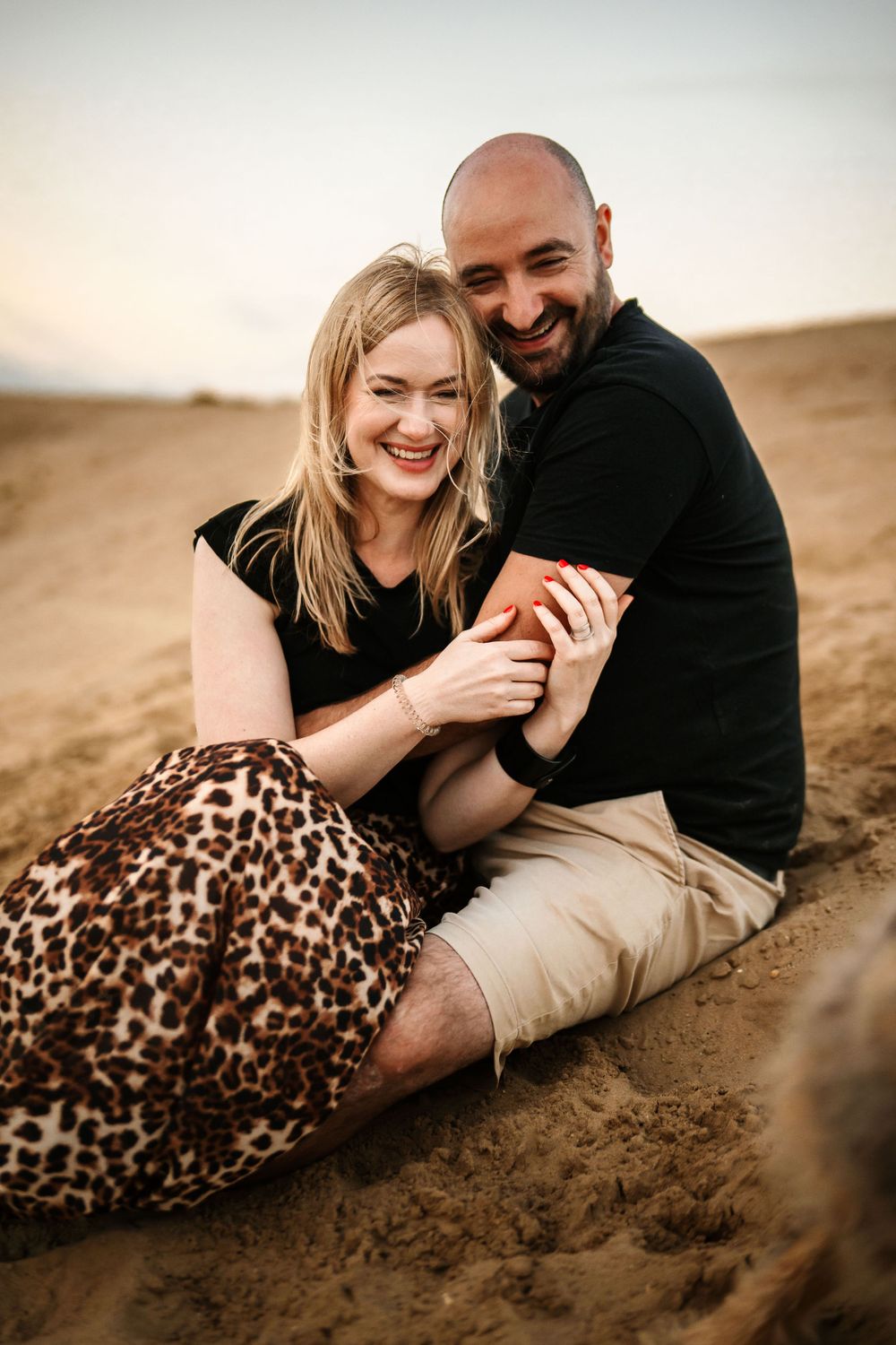 Happy couple laughing together on sand dunes during golden hour photoshoot.