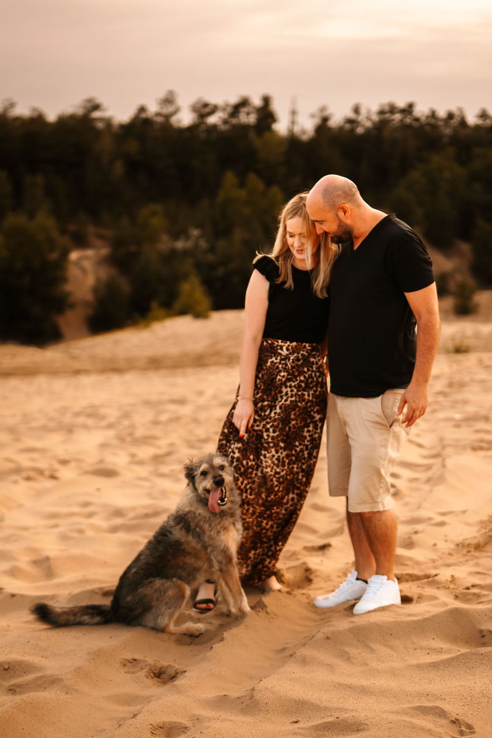 Happy couple with their scruffy dog posing on sand dunes at sunset.