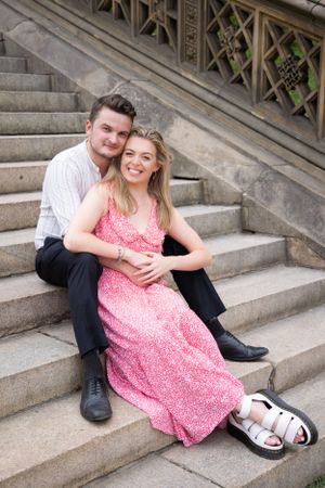 Smiling engaged couple sits together on stone steps at Bethesda Terrace, NYC.