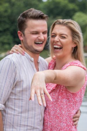 Joyful newly engaged couple laughs while woman shows off engagement ring outdoors.