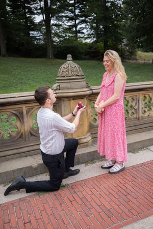 Man presents engagement ring to surprised woman in pink dress at Central Park.