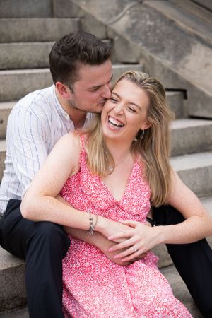Man kisses laughing woman on cheek while sitting on stone steps in park.