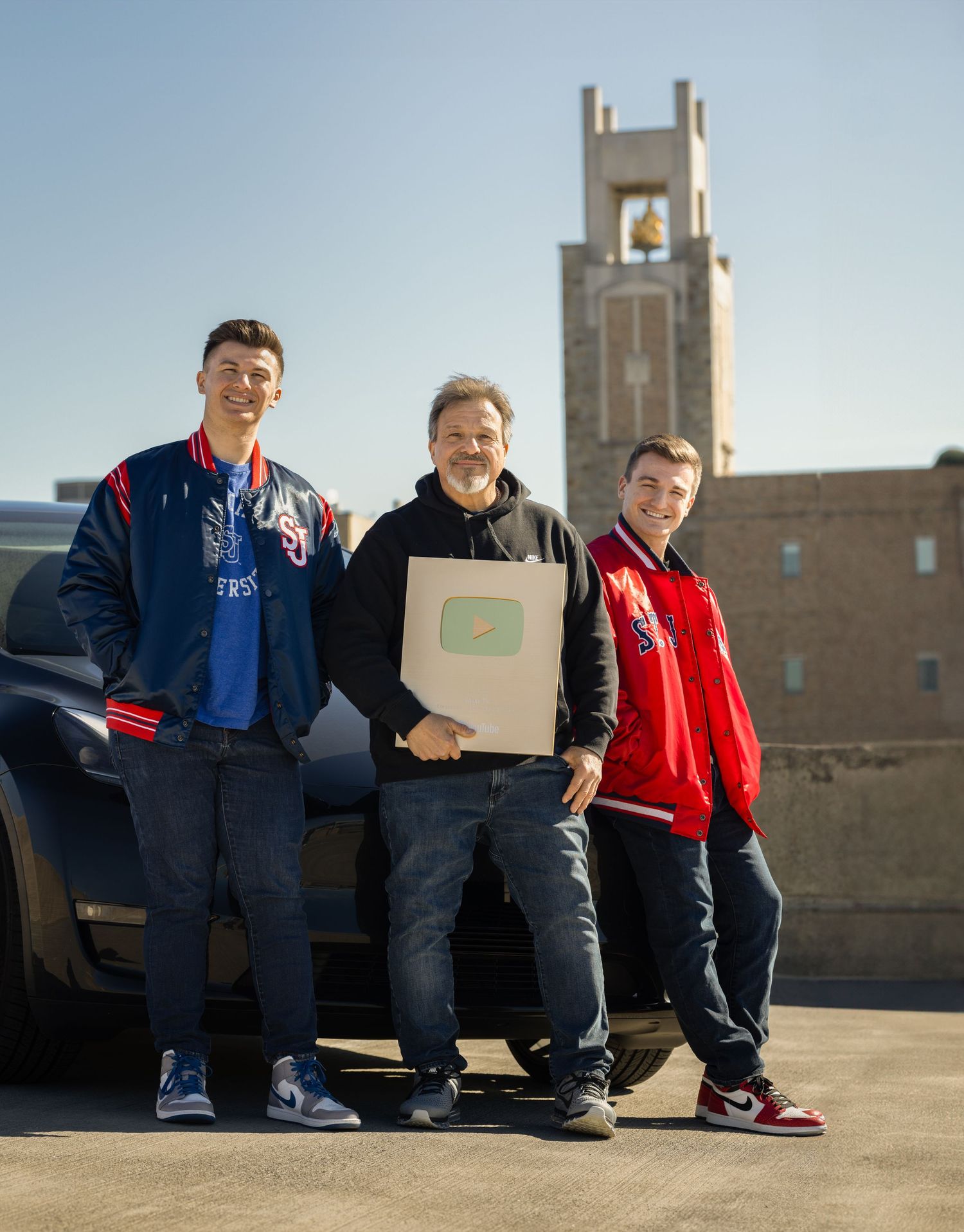 Three people standing by car with package in front of brick building with tower.