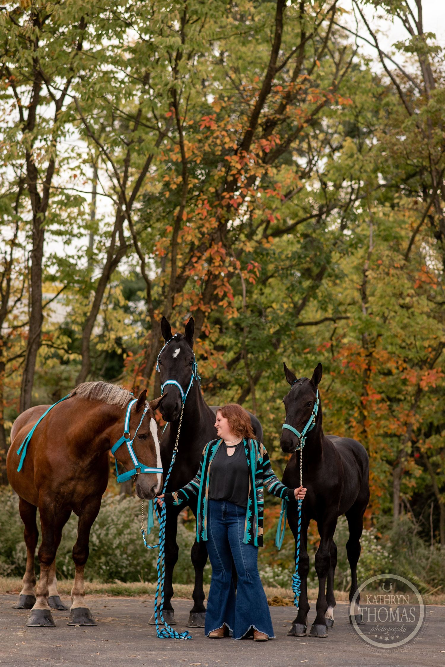 Handler poses with three horses wearing turquoise equipment against fall foliage.