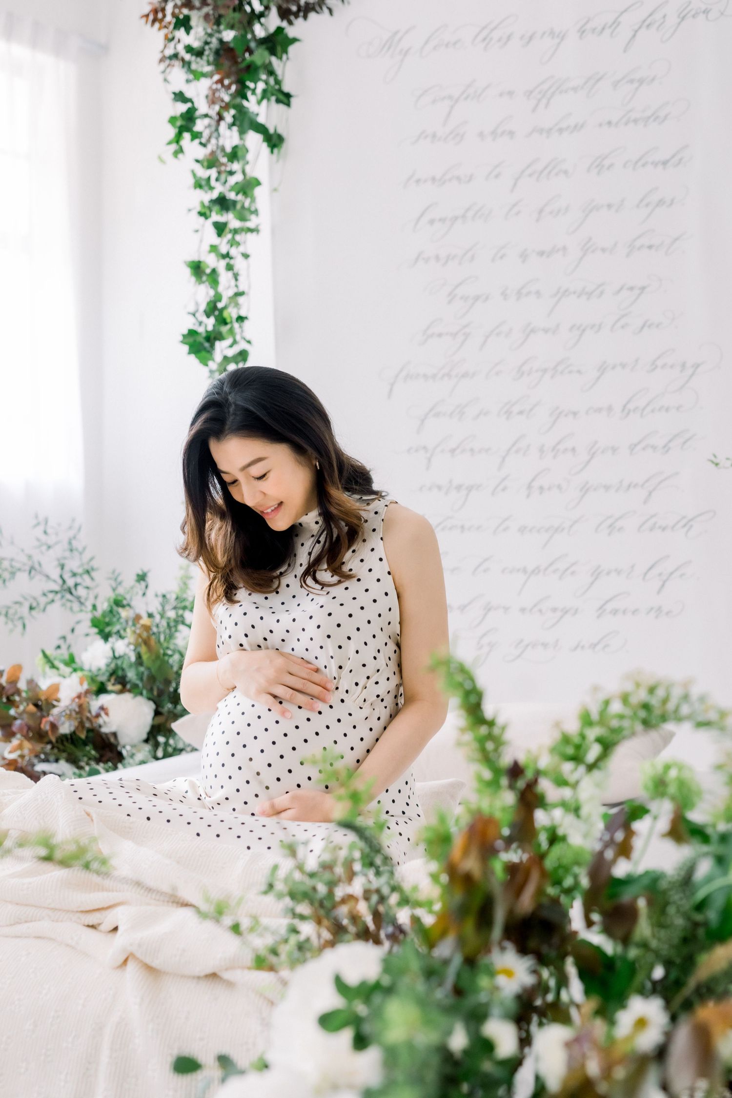 Expectant mother in polka dot dress sits serenely among floral decorations.