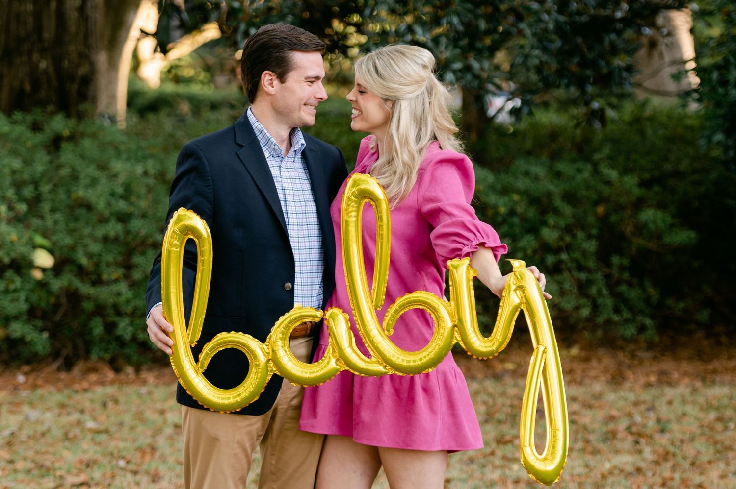 Happy couple holding a gold baby balloon script sign for a pregnancy announcement outdoors.