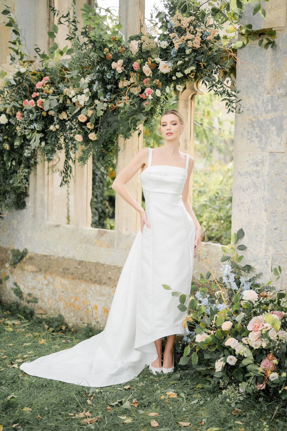 Wedding dress with train displayed in garden setting with pink roses and stone architecture.