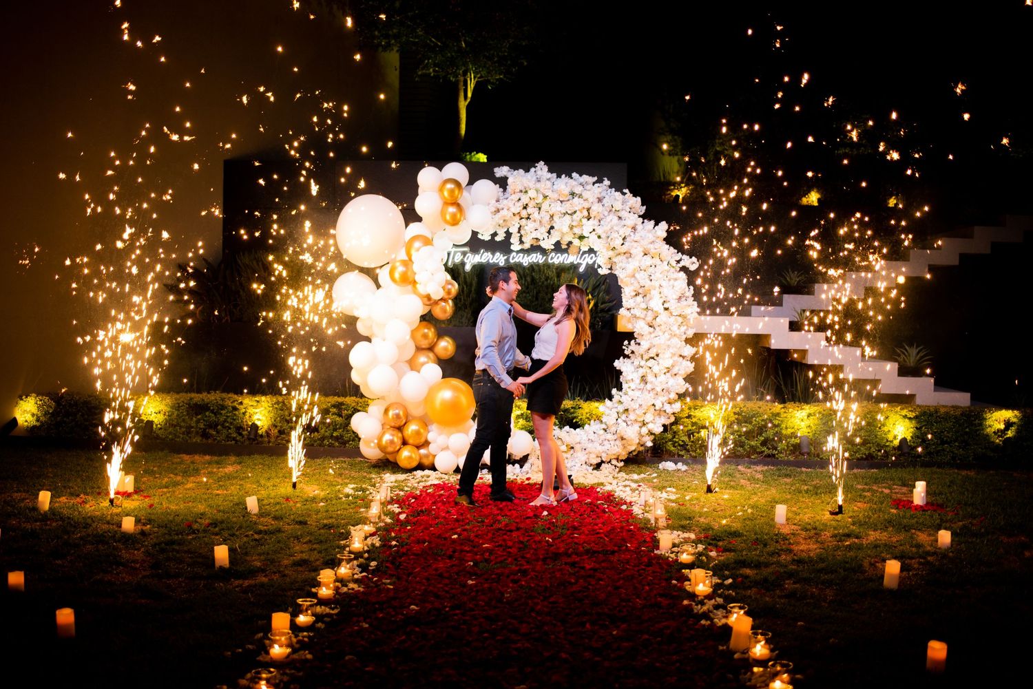 Romantic outdoor proposal setup with floral arch, rose petal aisle, balloons, and sparkling fireworks.