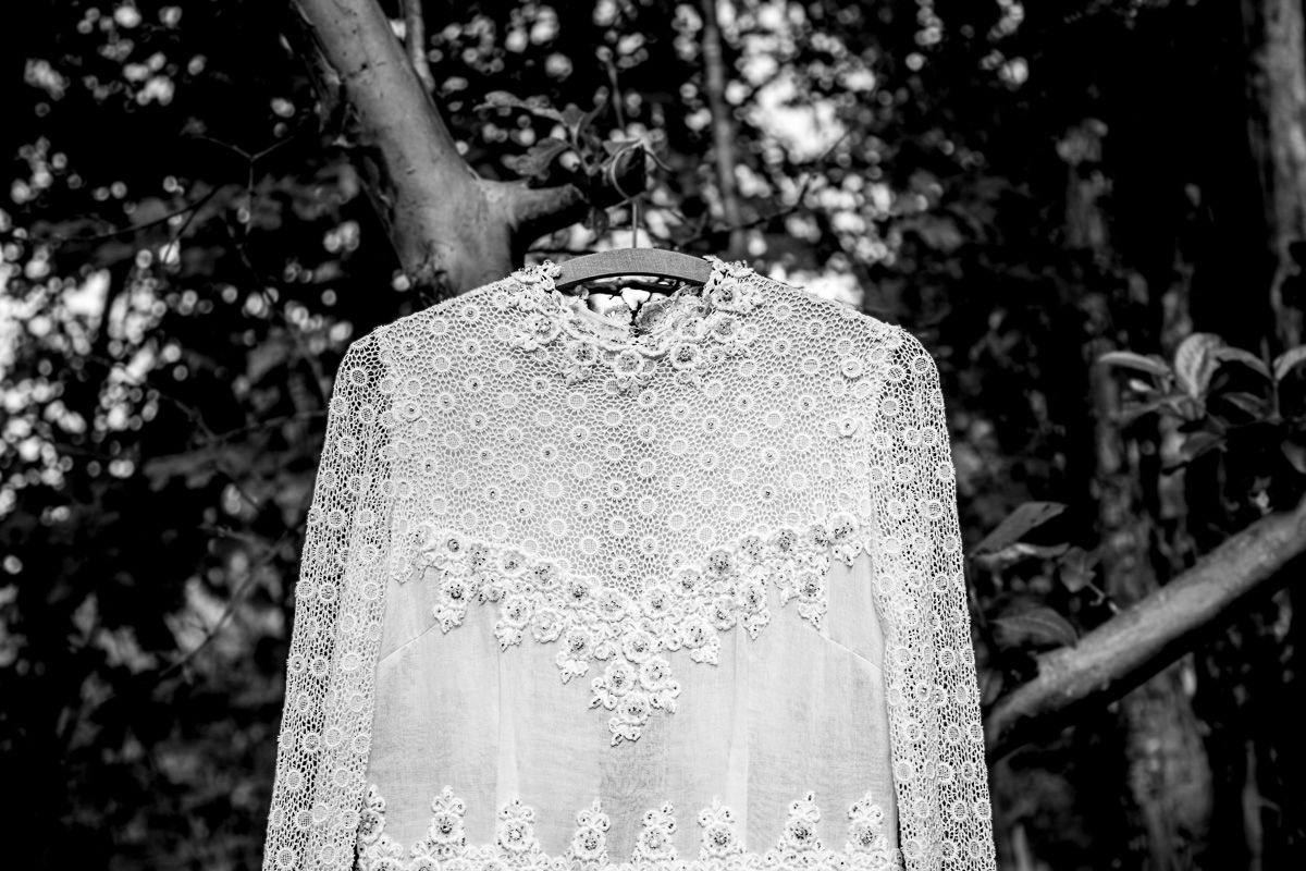 A delicate white lace wedding dress with intricate floral embroidery hangs from a hanger in a forest setting.