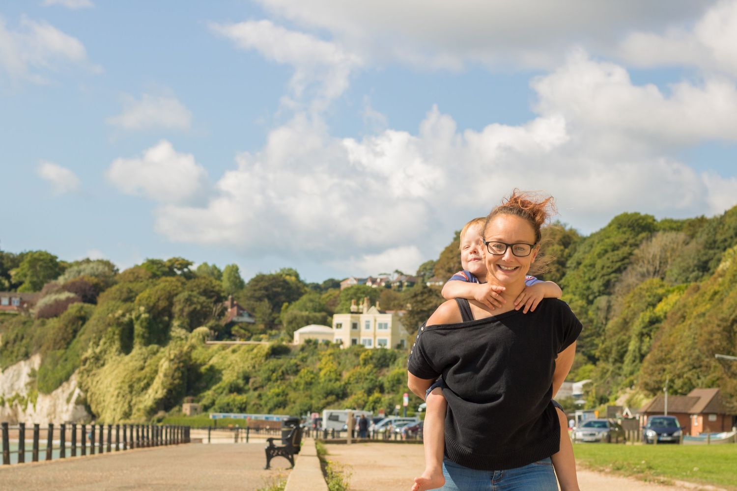 Photographer Kate Leach from Honeybees Photography walking along the beach in the sunshine with her son on her back