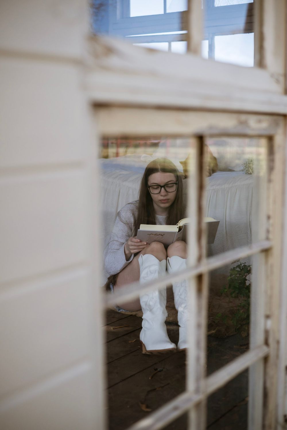 Person wearing glasses reads while sitting by a window in a cozy white indoor setting.