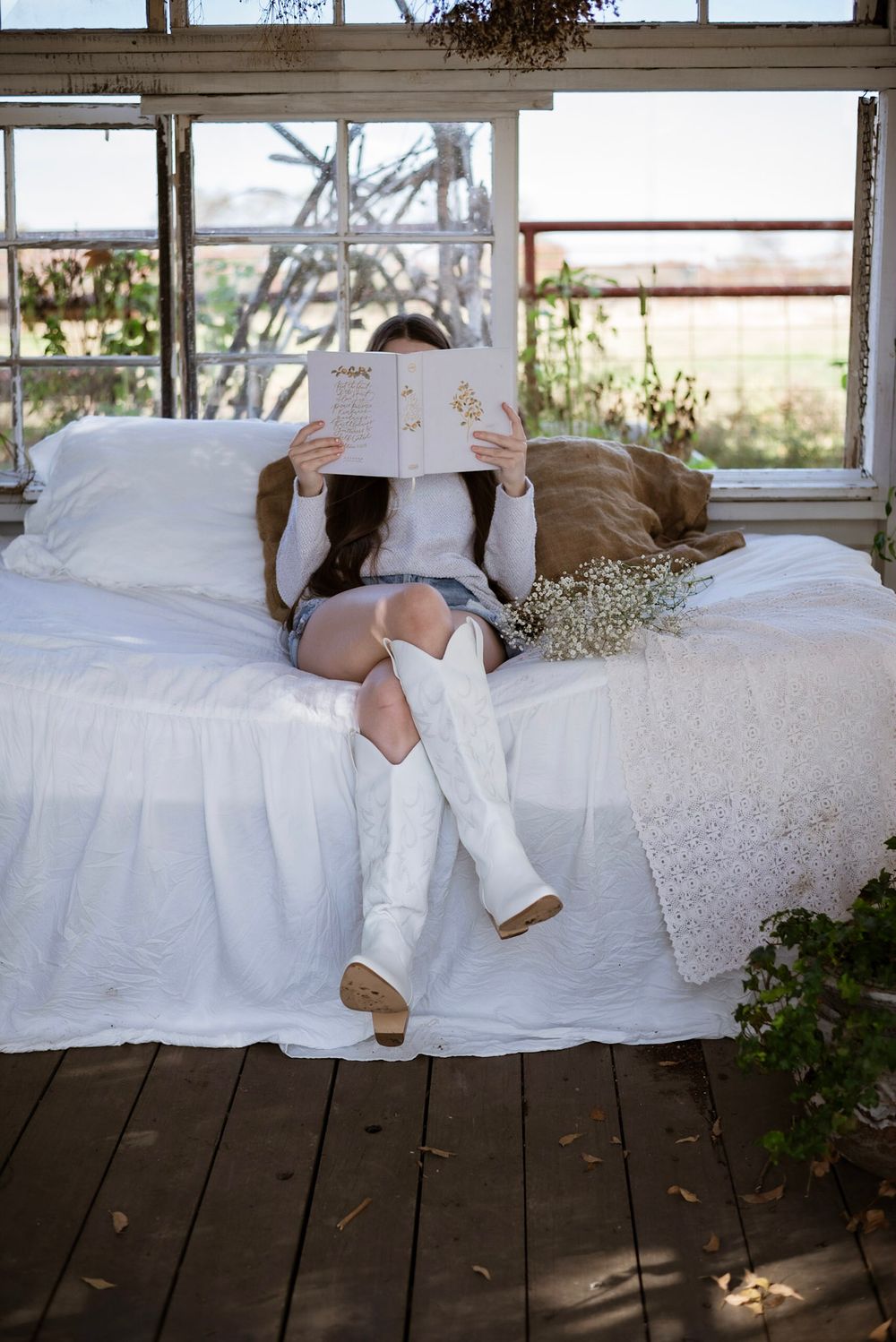 Person in white boots and denim shorts reads while relaxing on white bedding in sunroom.
