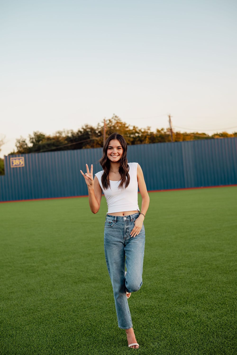 Stylish pose on baseball field wearing white top and jeans with natural sunset lighting.