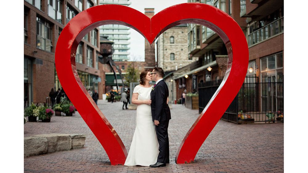 Couple in wedding attire pose inside large red heart sculpture on brick street.