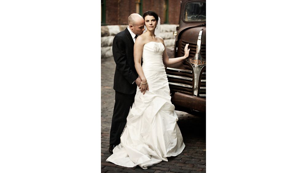 Elegant wedding couple posing next to vintage car with bride in flowing white gown.
