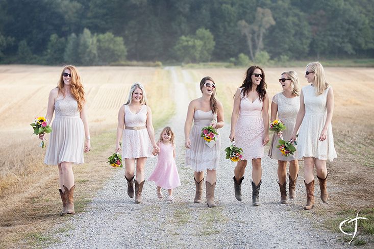 Bridesmaids in light pink dresses and cowboy boots walking together on a country path.