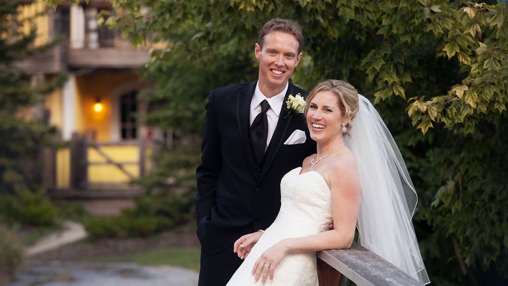 Wedding portrait of couple in formal attire standing in front of rustic building at dusk.