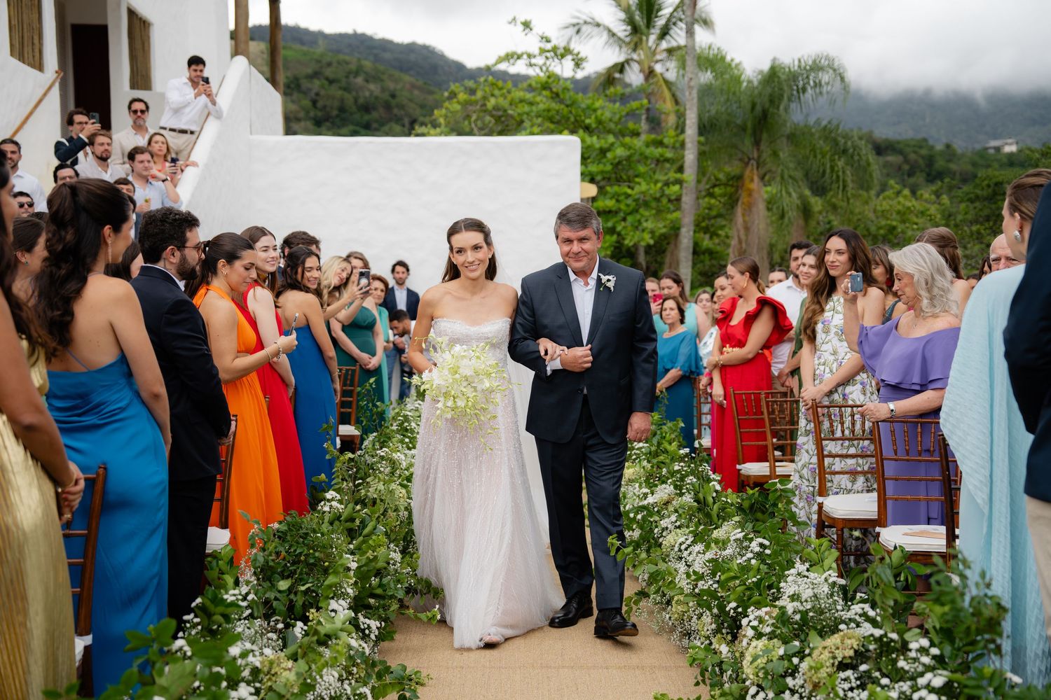 Wedding ceremony procession along garden path with colorfully dressed guests and tropical scenery.