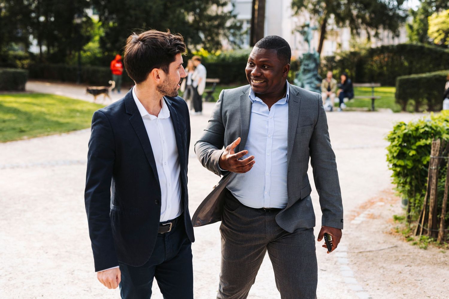 Two friends in suits walking and laughing together on garden path.