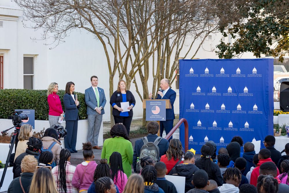 Crowd of students and attendees gather for outdoor presentation with speakers and blue backdrop.